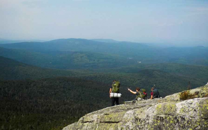 three people wearing backpacks rest on a rocky overlook with a green mountainous landscape in the background